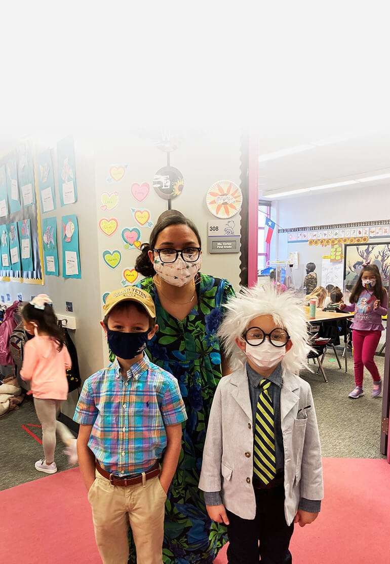 A student wears a messy white wig and lab coat, standing next to a classmate in a plaid top and baseball cap. Their teacher crouches behind them. All of them are wearing masks.