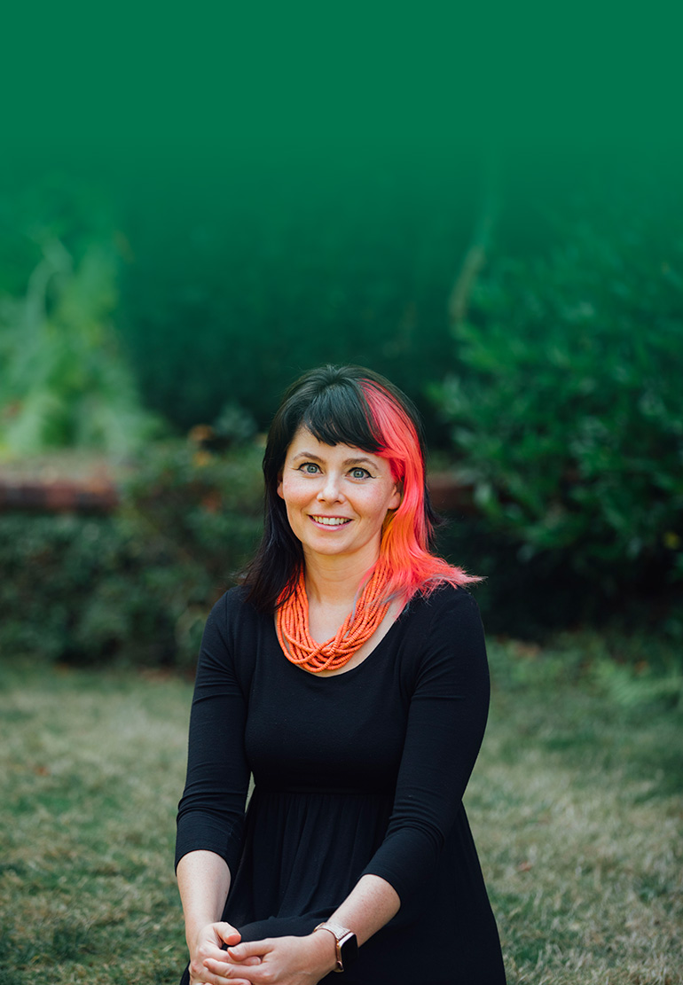 A women with black and pink hair smiles with greenery behind her.