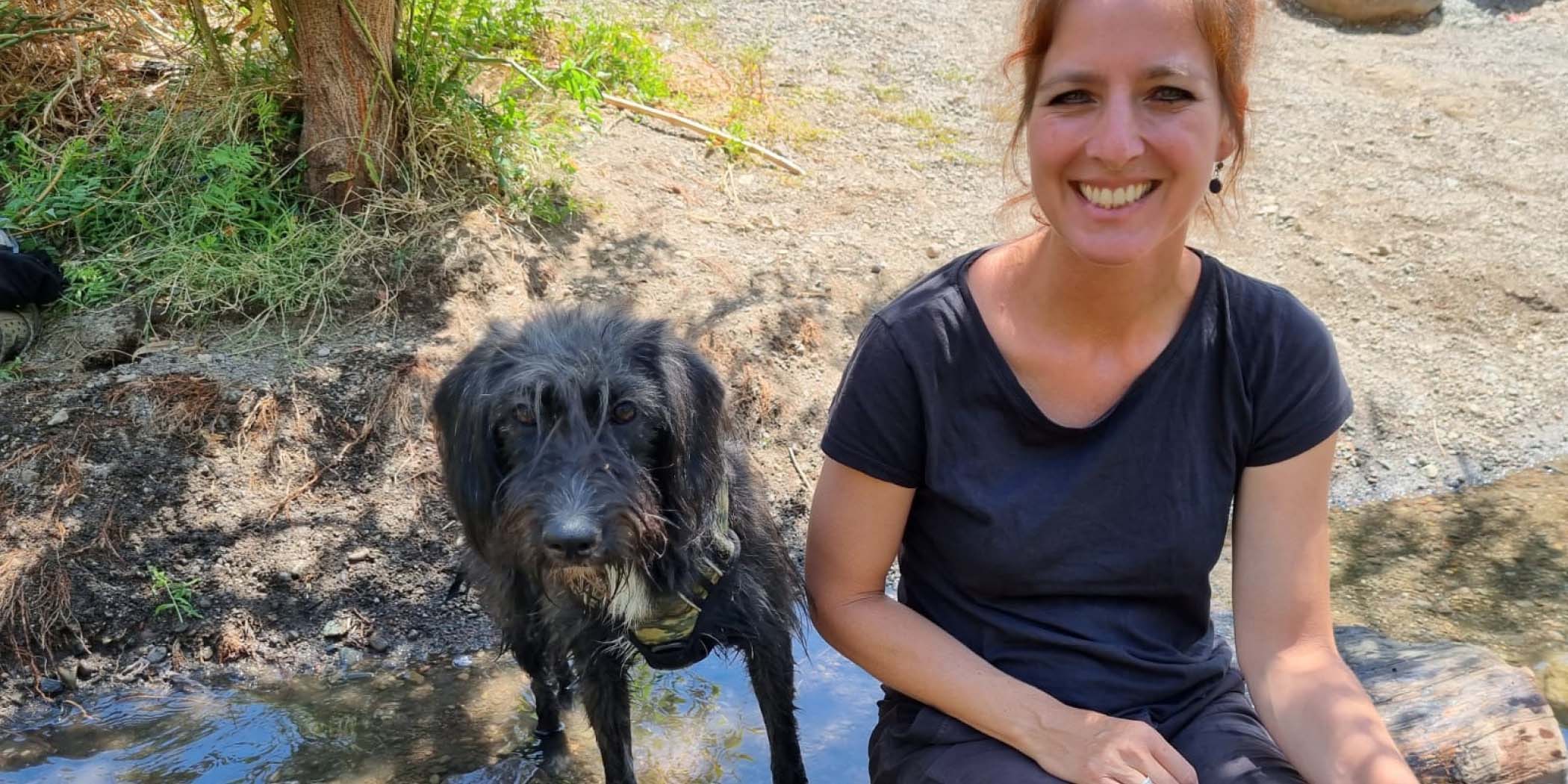 Alenka Lovy smiles and sits on a log outside next to a dog.
