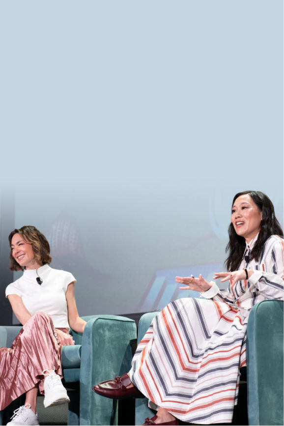 Priscilla Chan and Emily Chang speak on stage at SXSW. Priscilla gestures while talking, wearing a striped dress, and Emily smiles beside her, wearing a white top and pink pleated skirt.
